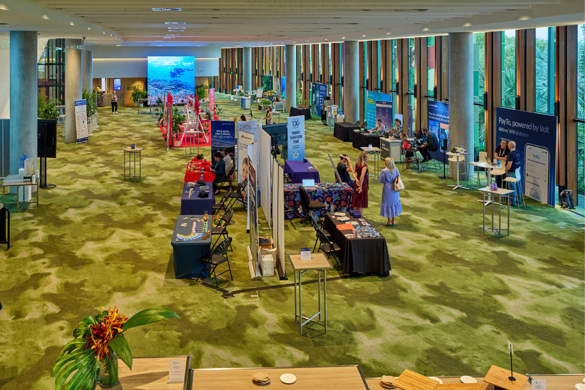 Large indoor space, Mezzanine Exhibition, with green-patterned carpet and high ceilings, featuring multiple booths and display tables arranged in rows. The Virgin Australia-branded activation area with a red carpet and white swing decorated with red flowers is visible in the background. A large digital screen at the far end displays an underwater scene with a sea turtle. The venue has tall glass windows along one side, allowing natural light to brighten the modern interior.