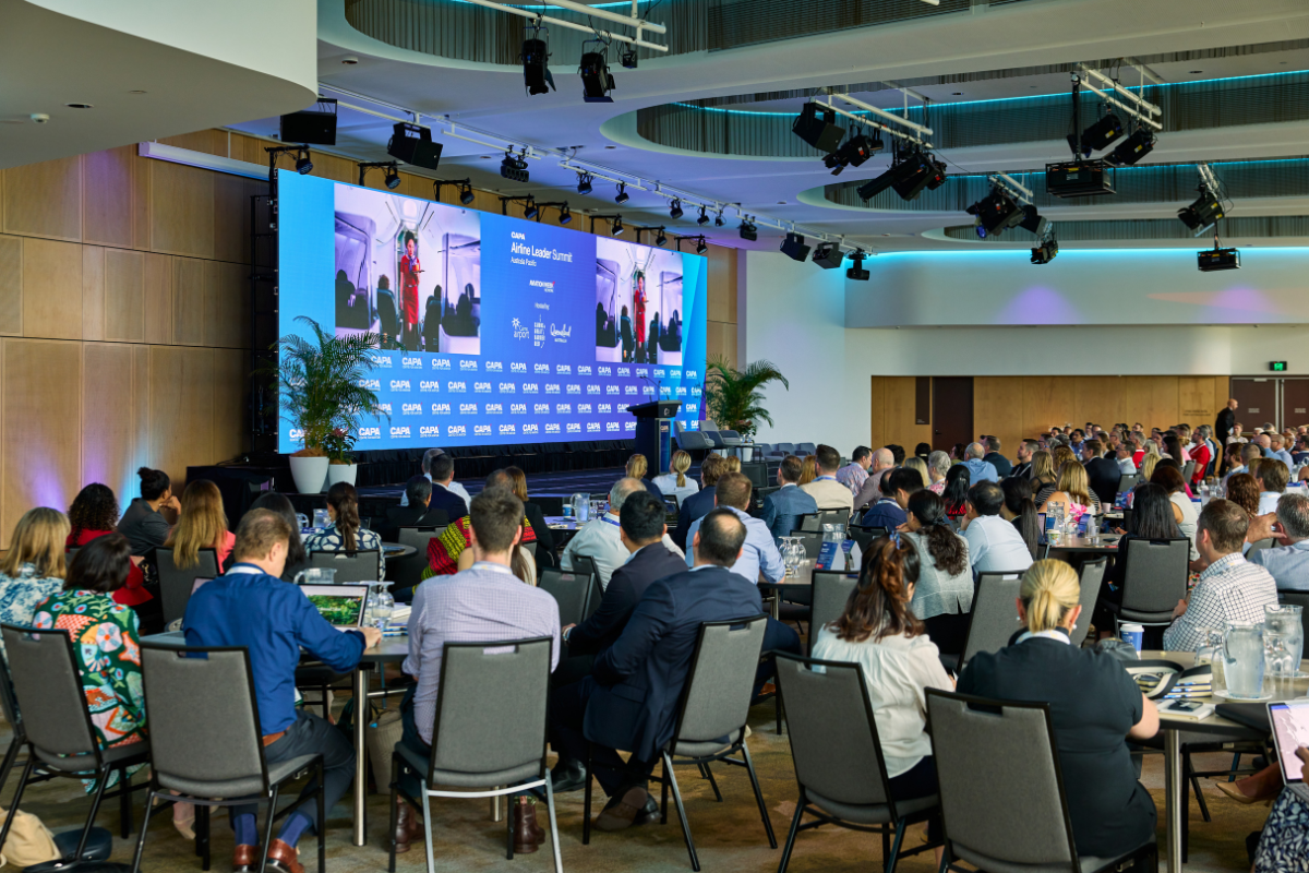 Conference room filled with attendees seated at round tables, facing a large stage with two big screens displaying a presentation titled “Aviation Leaders Summit.” The stage backdrop features a blue design with multiple sponsor logos, and two potted plants are placed on either side of the stage. The ceiling has modern lighting fixtures and black stage lights, creating a professional event setting.