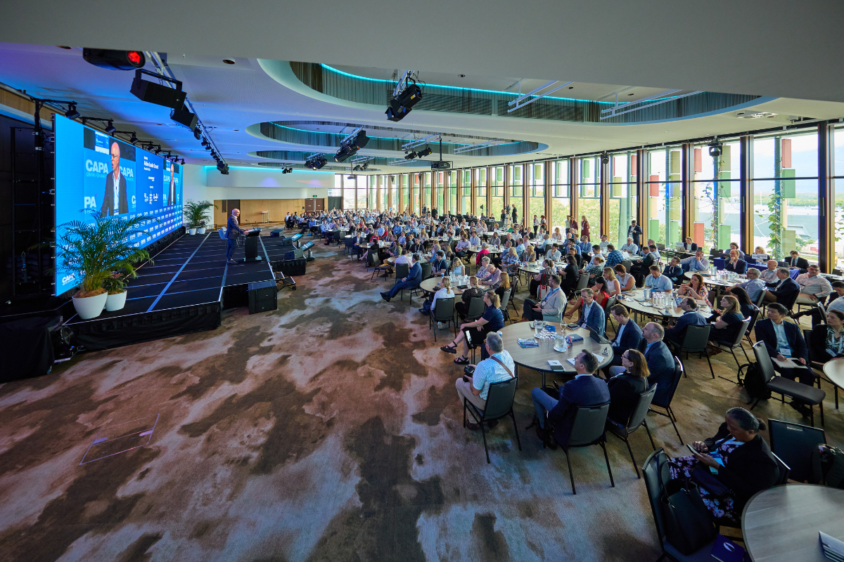 Large conference room, Trinity Room, with rows of round tables filled with attendees facing a stage. The stage features two large screens displaying a speaker and the event branding for “CAPA – Centre for Aviation.” The backdrop is blue with sponsor logos, and potted plants are placed on the stage. The room has floor-to-ceiling windows along one side, allowing natural light, and a modern ceiling with lighting fixtures and suspended stage lights.