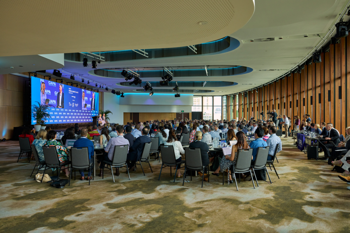 The Trinity Room filled with attendees seated at round tables, all facing a stage with two large screens displaying a presentation for the “CAPA – Centre for Aviation” event. The stage backdrop is blue with sponsor logos, and potted plants are placed on either side. The room features a modern ceiling with lighting fixtures and stage lights, and a wall of tall windows on the right side allowing natural light into the space.