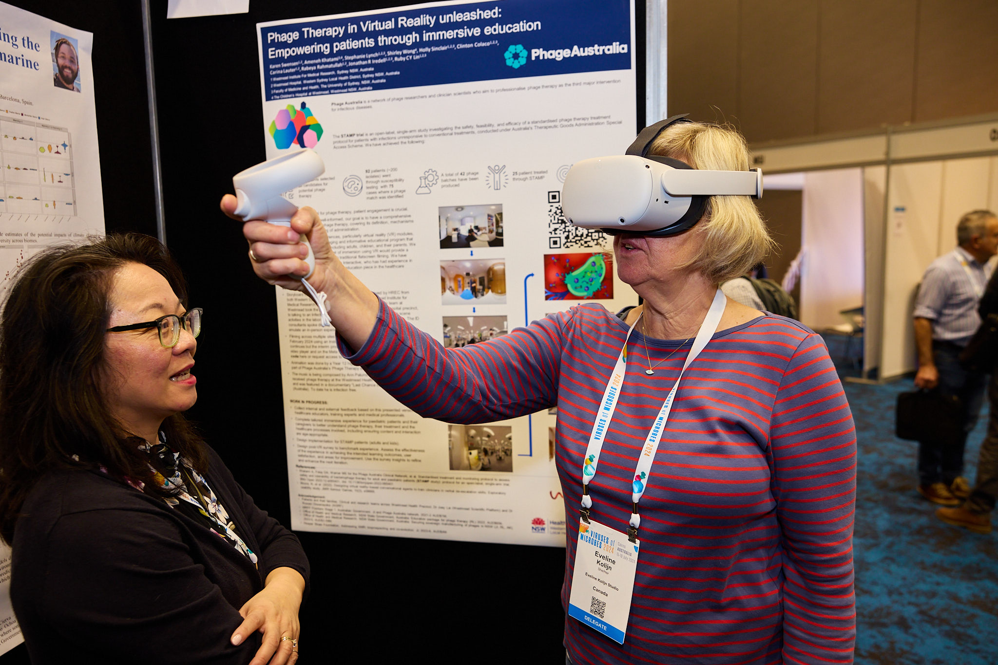 Person wearing a virtual reality headset and holding a VR controller while interacting with a display at a conference. Behind them is a poster titled ‘Phage Therapy in Virtual Reality unleashed: Empowering patients through immersive education,’ featuring text, images, and a QR code. Another person stands nearby observing