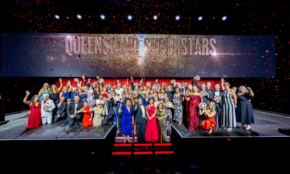 Large group of Queensland Tourism Awards winners celebrating on stage, holding circular trophies, with red confetti falling and a backdrop displaying the words “Queensland Superstars.”