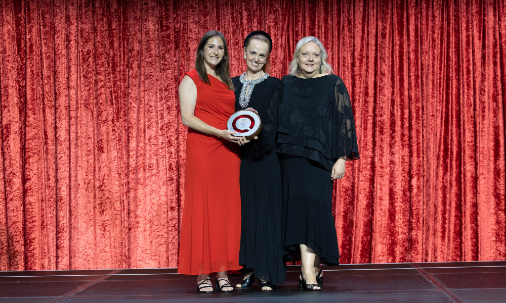 Melissa Pringle, Marketing Manager at Cairns Convention Centre; Elizabeth Hackett, Chair of QTA Judges; and Janet Hamilton, General Manager at Cairns Convention Centre, standing on stage holding a Queensland Tourism Award trophy in front of a red curtain backdrop.