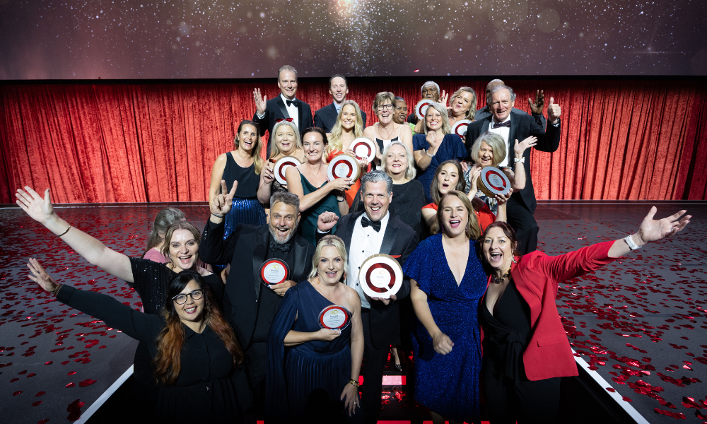 Group of award recipients posing on stage at the Queensland Tourism Awards, holding circular trophies with a red and white design, against a backdrop of red curtains and a starry display.