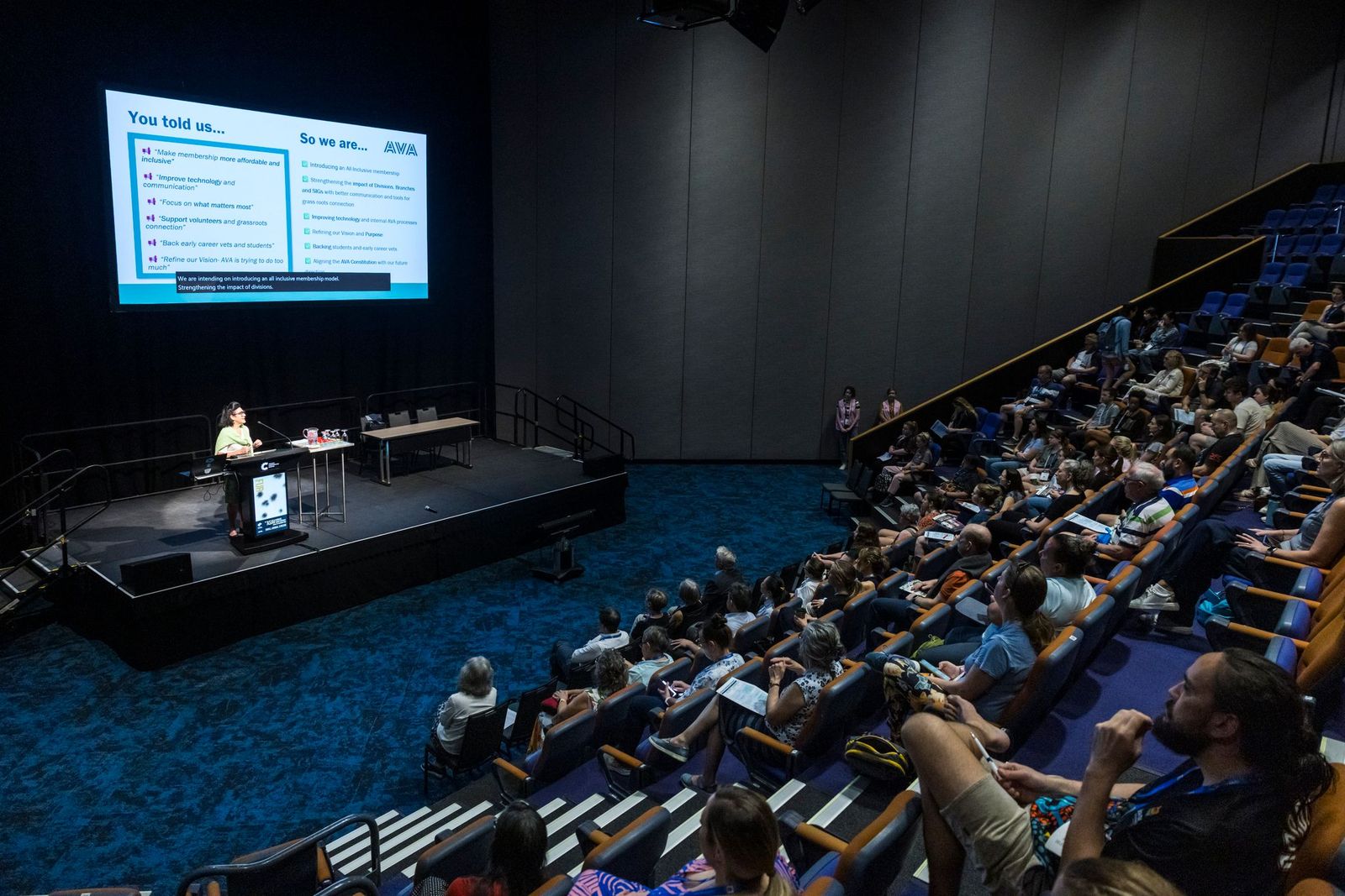 Conference session in Auditorium at ASAV 2025 with a speaker presenting on stage in front of a large screen displaying key points. Audience members are seated in tiered rows, attentively watching the presentation in a modern lecture hall.
