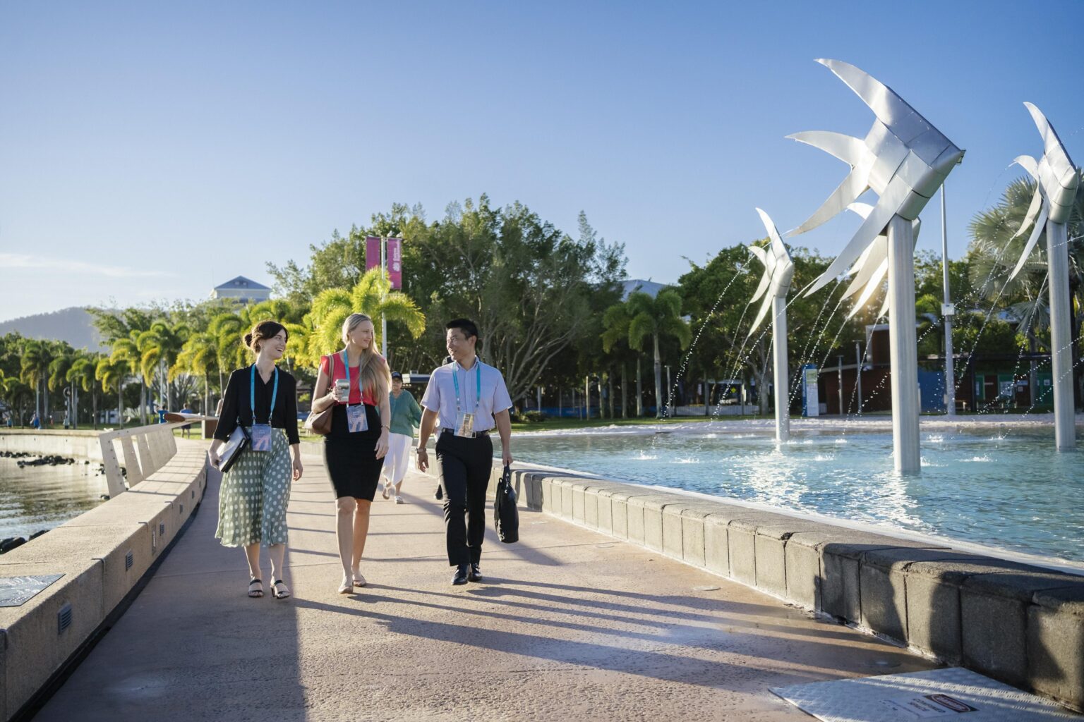 Three conference delegates walk along the Cairns Esplanade near the Cairns Lagoon. They are on a wide paved pathway beside a large water feature with silver fish sculptures and fountains. Palm trees and greenery line the background under a clear blue sky, creating a tropical setting.
