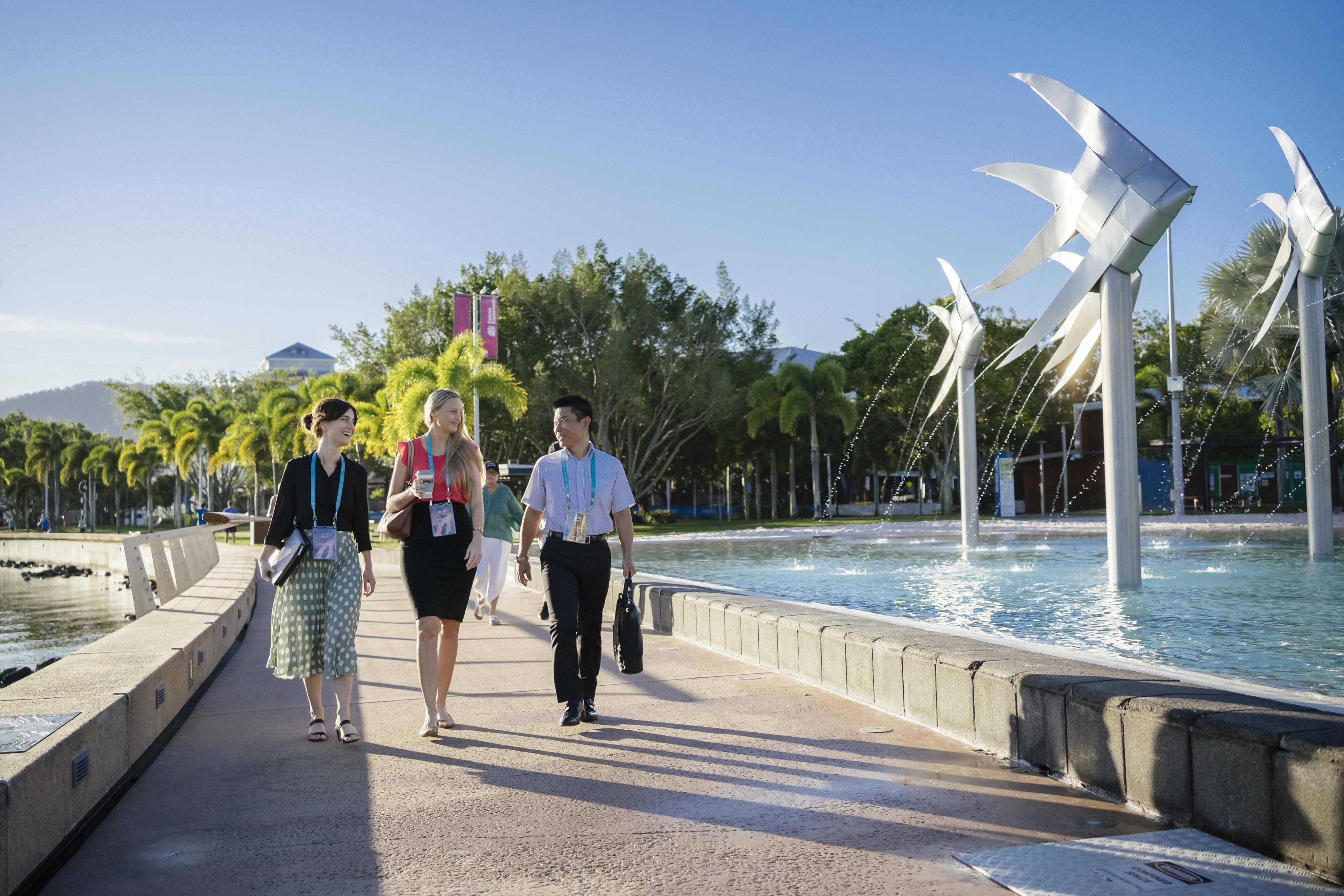 Three conference delegates walk along the Cairns Esplanade near the Cairns Lagoon. They are on a wide paved pathway beside a large water feature with silver fish sculptures and fountains. Palm trees and greenery line the background under a clear blue sky, creating a tropical setting.
