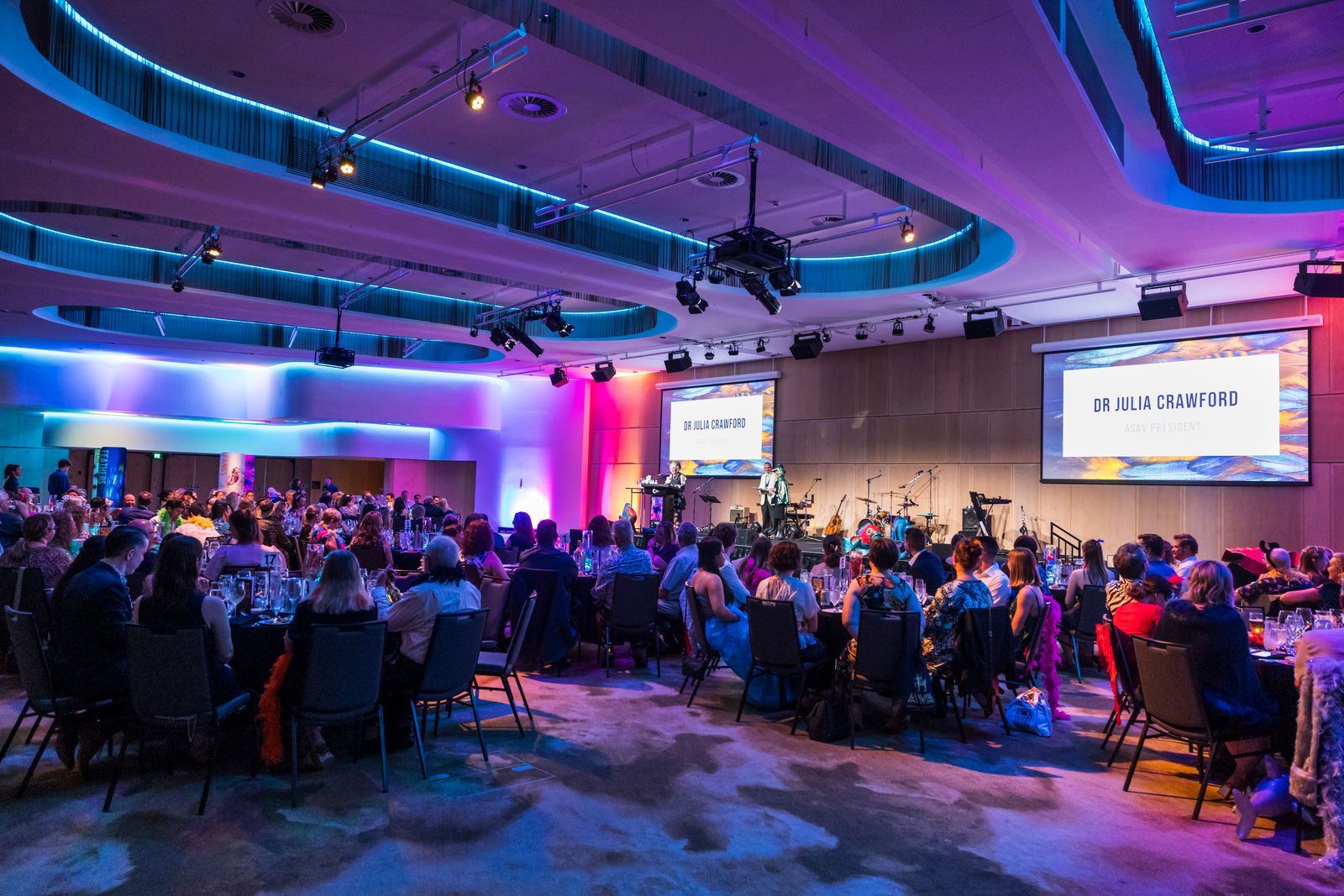 ASAV 2025 Gala Dinner in Trinity Room, a spacious area with elegant lighting in blue and purple tones. Guests are seated at round tables enjoying the event, while a speaker addresses the audience from a stage with two large screens displaying the name 'Dr Julia Crawford.' Musical instruments are set up on stage for entertainment.