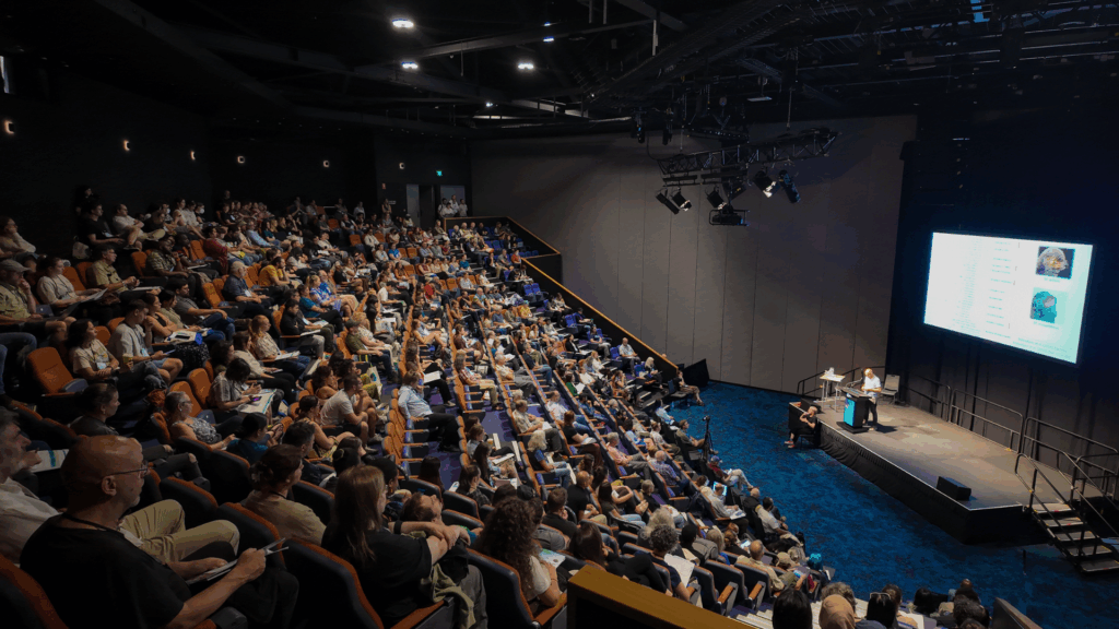 Large auditorium filled with delegates attending a plenary session at the International Bat Research Conference. The audience is seated in tiered rows facing a stage where a presenter stands at a podium next to a large screen displaying slides with text and images related to bat research.