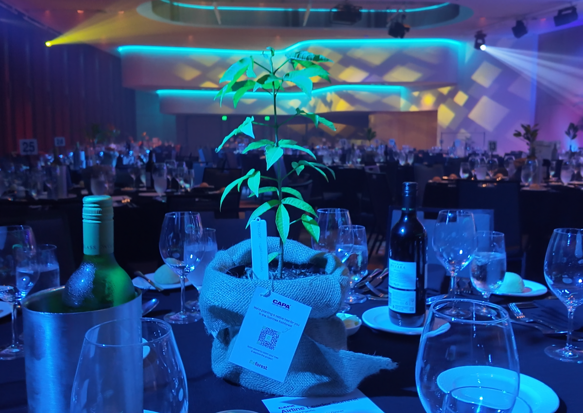 A decorated gala dinner table featuring a small potted tree as a centrepiece, surrounded by glassware, plates, and bottles of beverages, with a softly lit event stage and ambient blue lighting in the background.