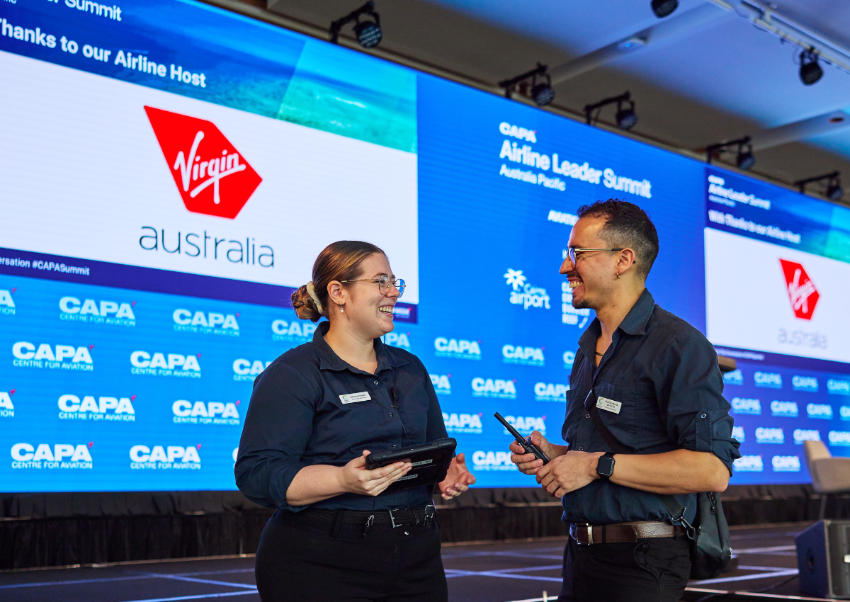 Two Cairns Convention Centre audio‑visual staff members stand in front of a large LED screen at the CAPA Airline Leader Summit, discussing technical setup while holding electronic devices.