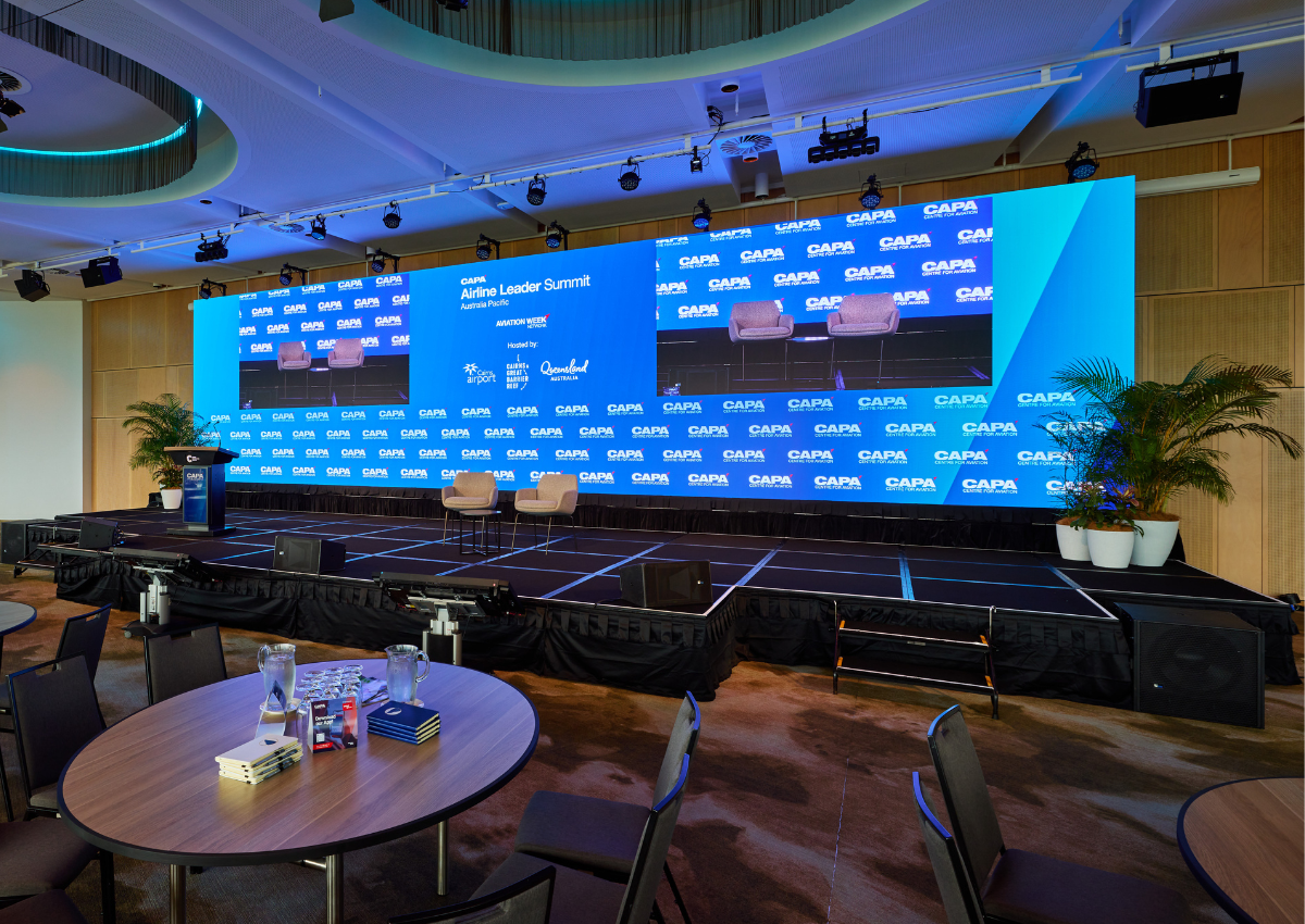 A stage setup for the CAPA Airline Leader Summit featuring a large LED screen with event branding, two chairs arranged for a panel discussion, and round tables set for delegates inside the Trinity Room at Cairns Convention Centre.