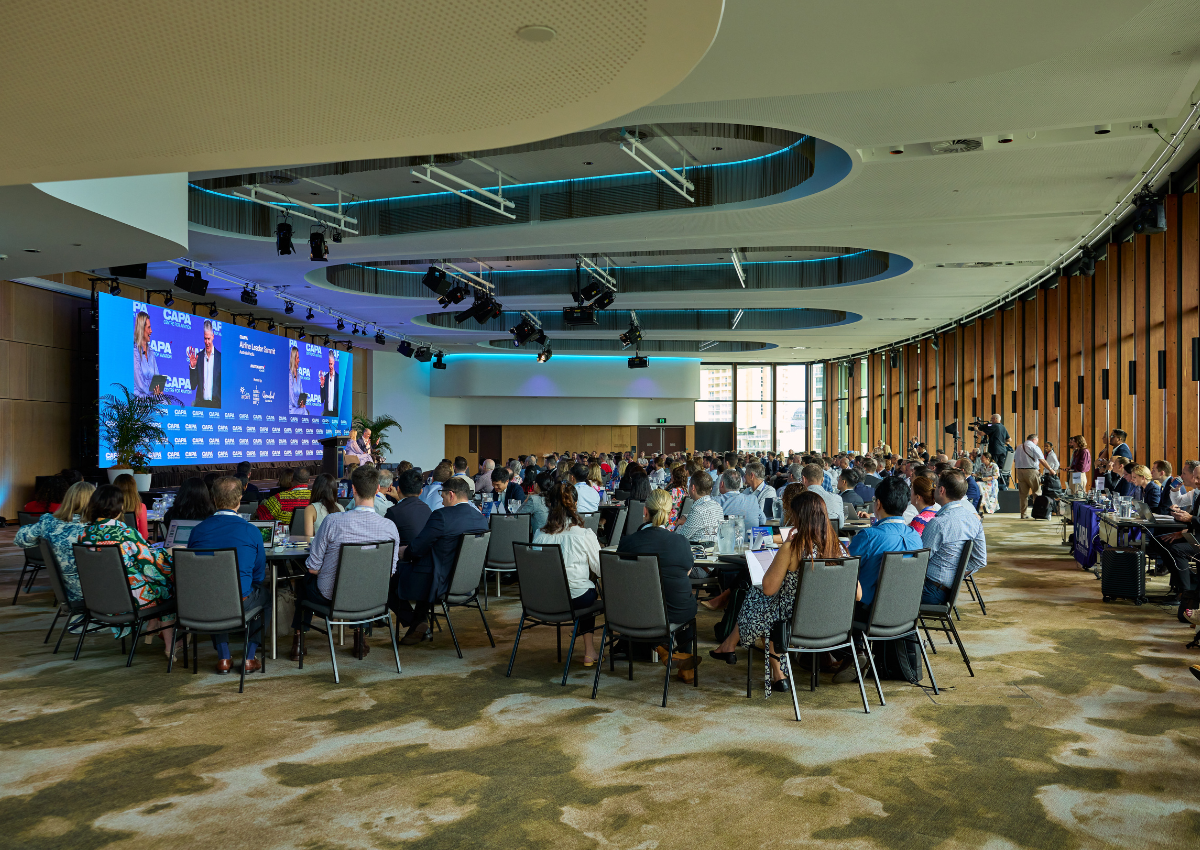 A large audience in the Trinity Room at Cairns Convention Centre seated at round tables listens to a speaker on stage at the CAPA Airline Leader Summit, with a wide LED screen displaying event branding inside a modern, spacious conference room at the Cairns Convention Centre.