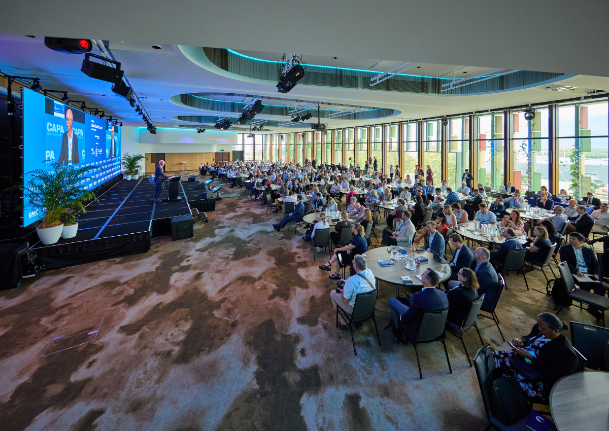 A large audience in the Trinity Room at Cairns Convention Centre seated at round tables listening to a speaker on stage at the CAPA Airline Leader Summit, with large LED screens displaying event branding inside a bright, modern conference hall with floor‑to‑ceiling windows.