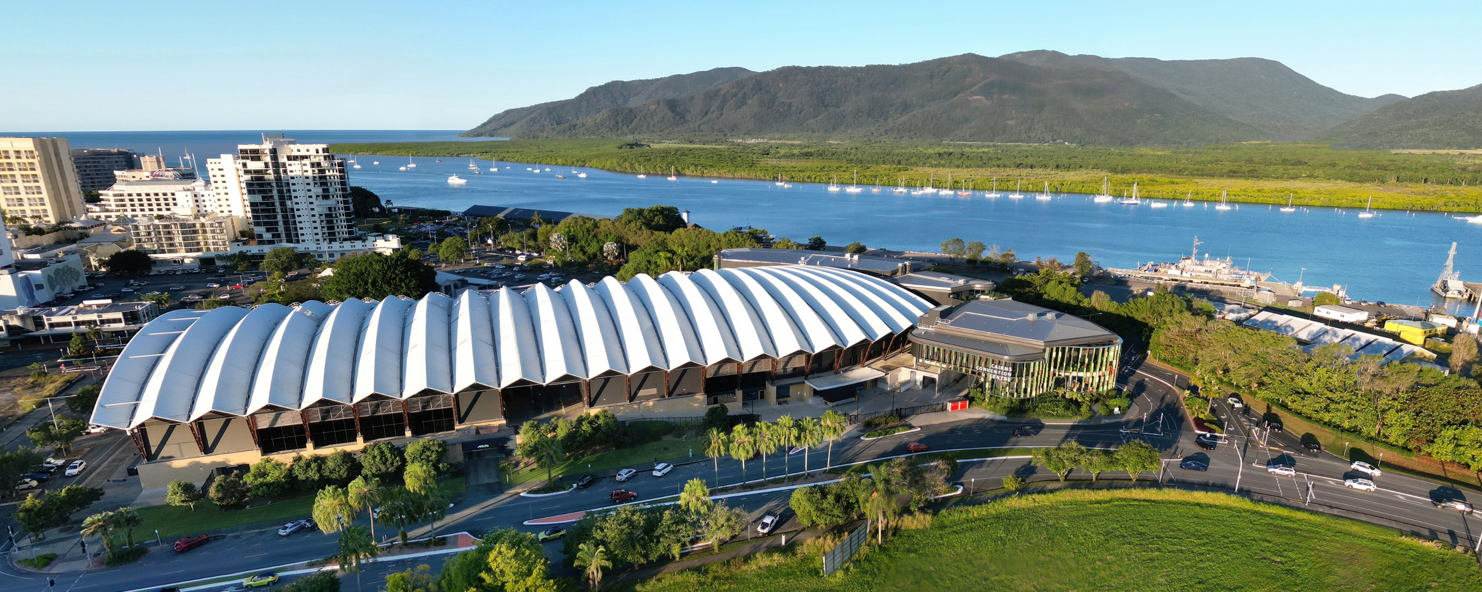 Aerial view of the Cairns Convention Centre with its distinctive white, wavy roof, surrounded by urban buildings and green spaces. In the background, the Trinity Inlet is visible with boats docked along the shore, and beyond it, the lush Wet Tropics Rainforest and distant mountains under a clear blue sky.