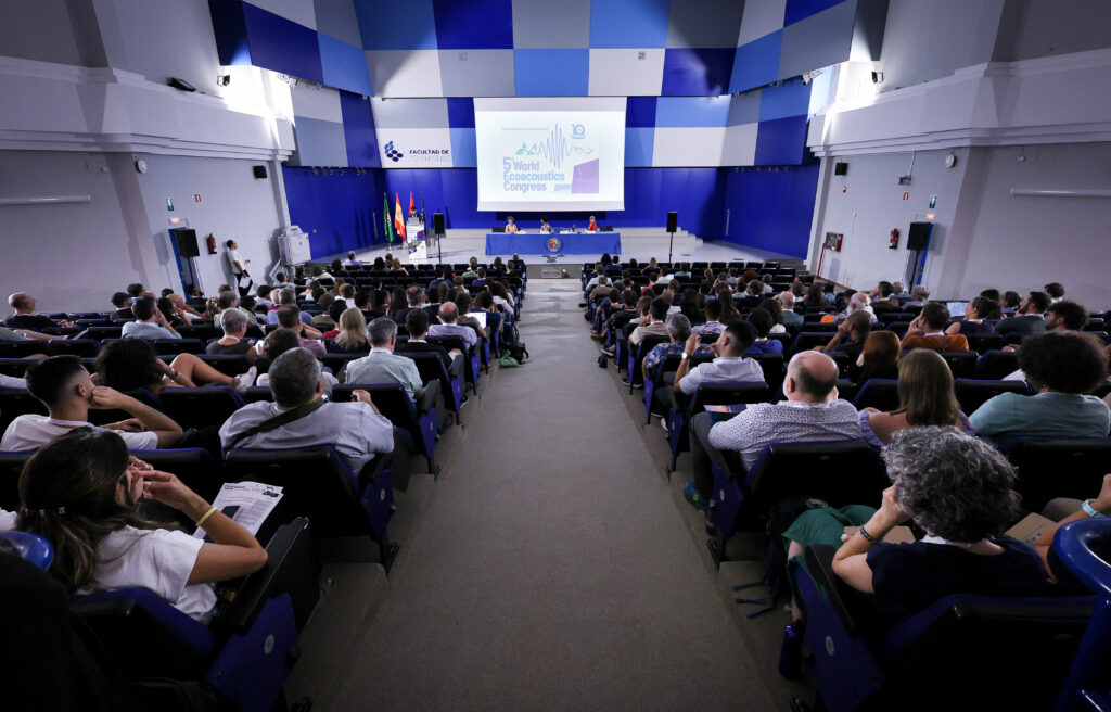 Wide view of a large lecture-style auditorium filled with seated attendees facing a stage, where a panel sits at a long table beneath a projection screen displaying ‘5th World Ecoacoustics Congress
