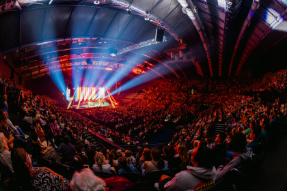 A wide-angle view of a large audience watching a performance on a brightly lit stage in a concert hall. Blue and red spotlights illuminate the stage and cast beams across the darkened arena filled with spectators.