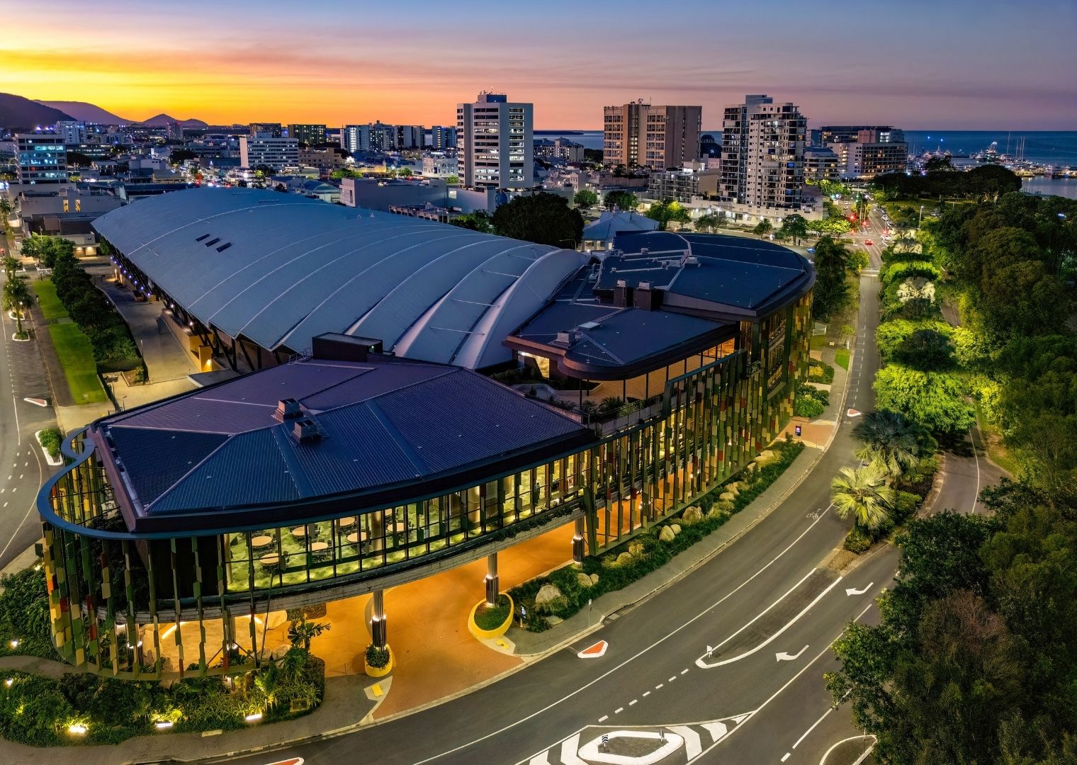 Aerial view of the Cairns Convention Centre with its curved, glass-accented architecture, surrounded by roads and greenery, with city buildings and the sea visible at sunset.